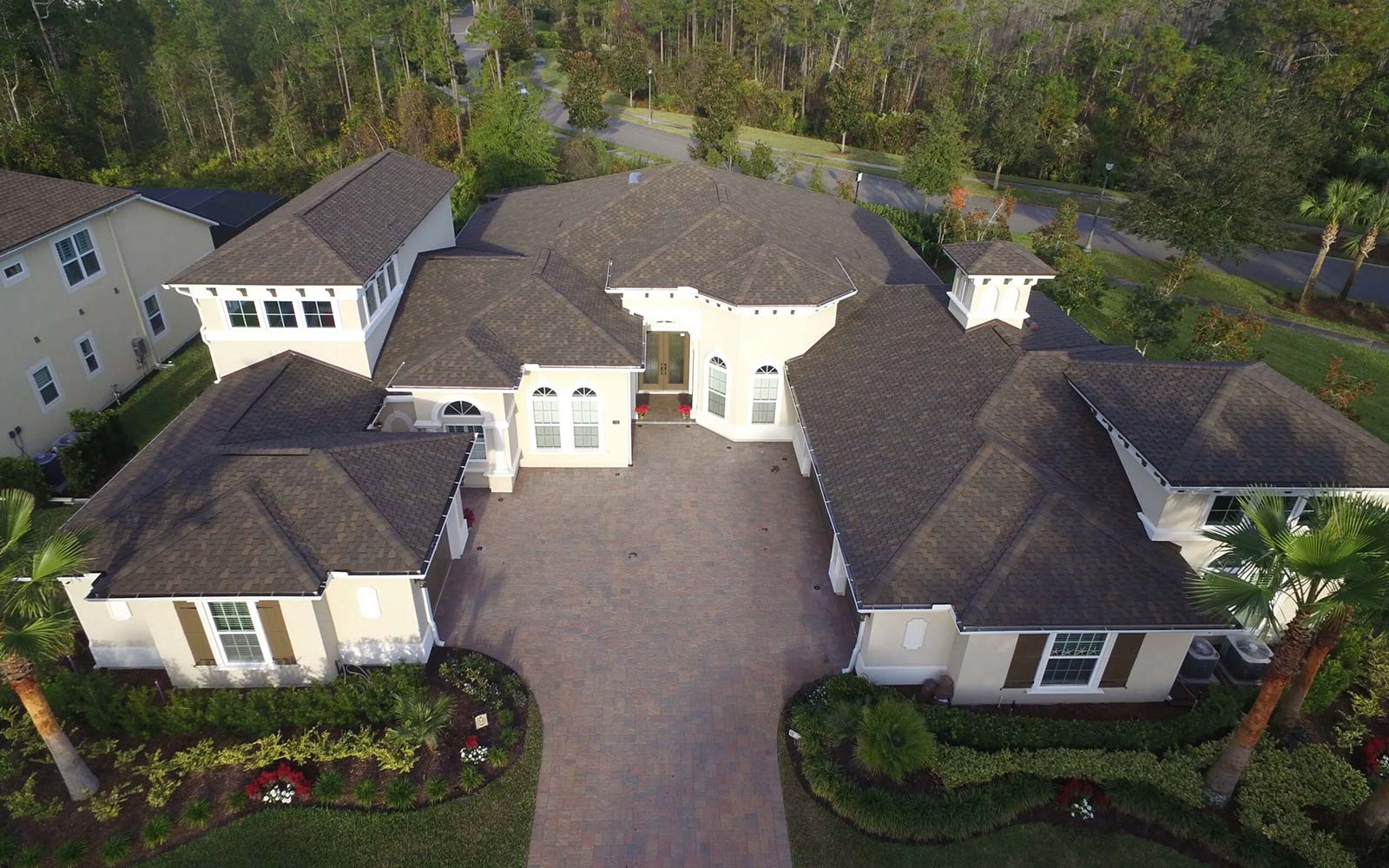 Aerial view of a large single-story home with a brown shingle roof, paved driveway, and landscaped front yard bordered by trees.