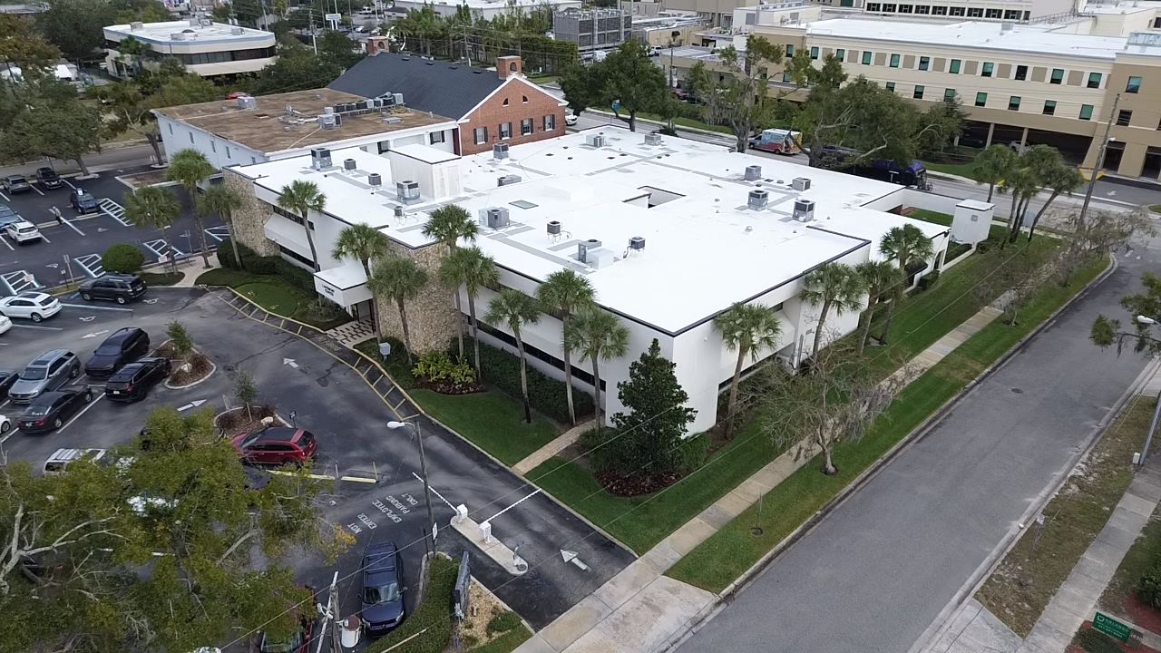 Aerial view of a large commercial building with a bright white flat roof, multiple rooftop HVAC units, palm trees along the perimeter, adjacent parking areas, and surrounding office buildings and streets.
