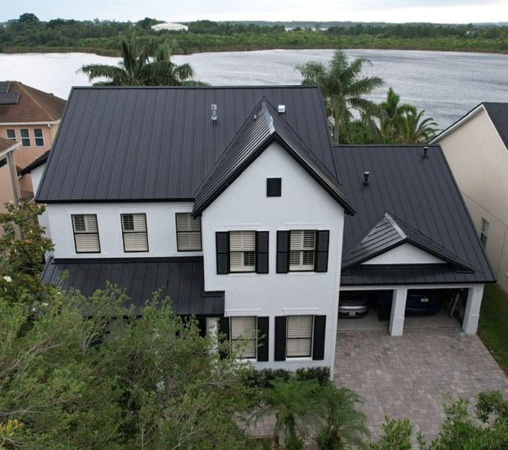 Aerial view of a Florida waterfront home with a black standing seam metal roof, showing steep rooflines, dormers, garage area, and lakefront surroundings.