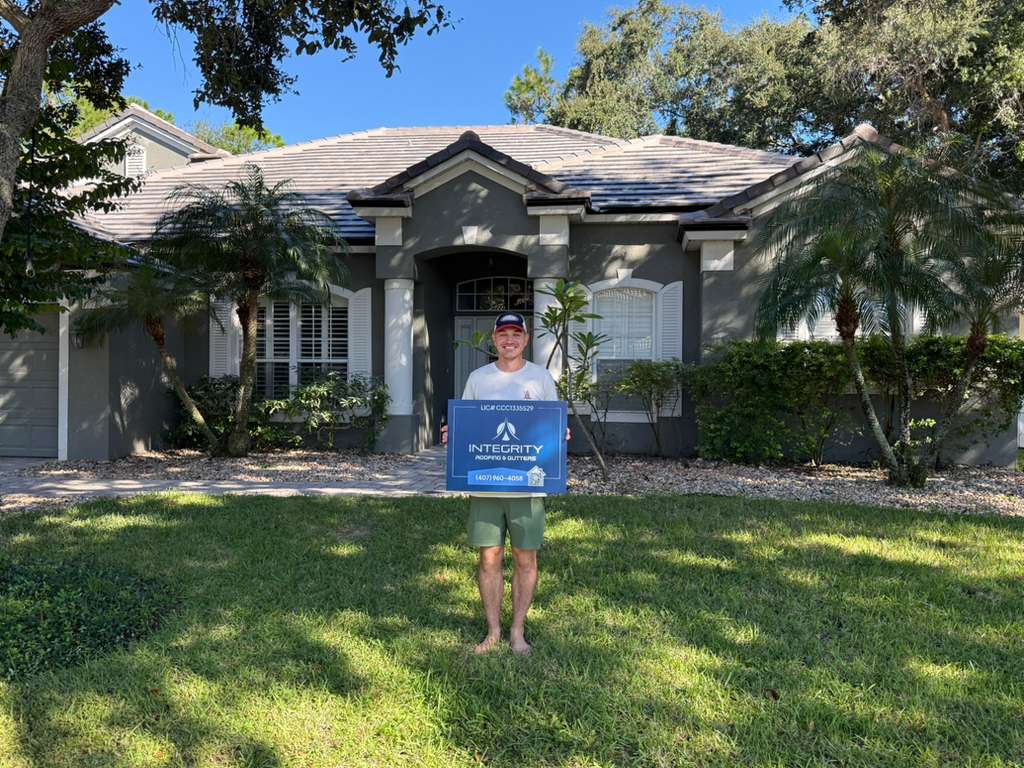 Homeowner standing barefoot on a green lawn in front of a single-story gray stucco house with a light-colored tile roof, holding an Integrity Roofing & Gutters sign, with palm trees and landscaping visible under a clear blue sky.