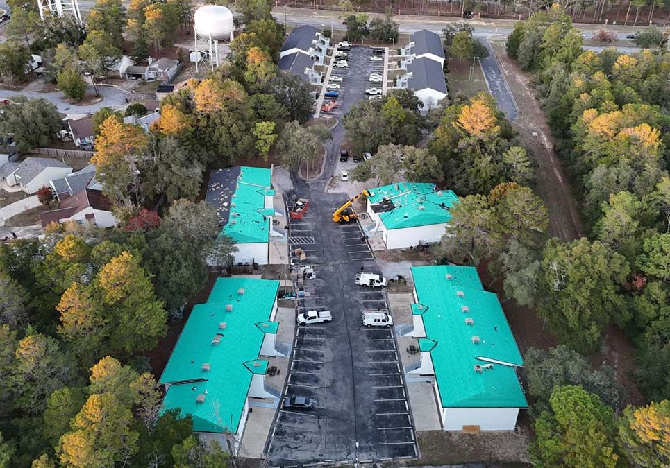 Aerial view of a small apartment or townhouse complex surrounded by trees, featuring multiple low-rise buildings with teal roofing under installation, parked work vehicles, construction equipment, and a central paved driveway with parking spaces.