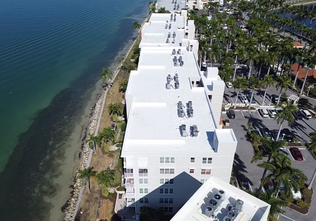 Aerial view of a long waterfront residential building with a bright white flat roof, multiple rooftop HVAC units, palm-lined parking areas, and clear blue-green water running along the shoreline beside the property.