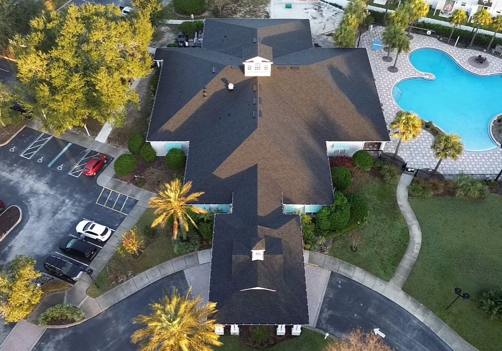 Aerial view of a residential clubhouse building with a dark shingle roof, adjacent parking lot, palm trees, landscaped walkways, and a resort-style swimming pool on the right side.