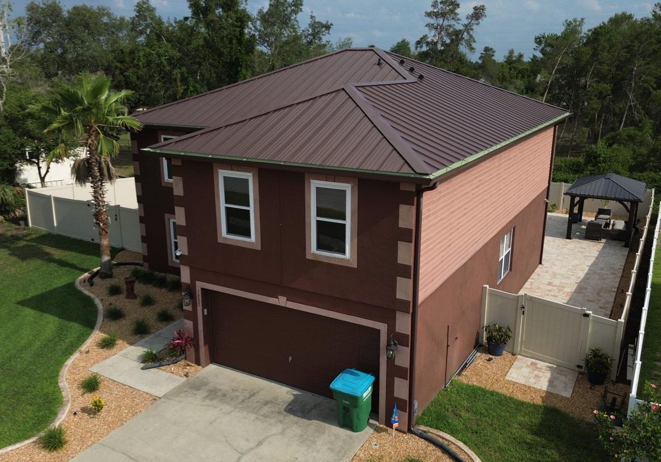 Two-story brown stucco home with a dark brown standing-seam metal roof, viewed from above, featuring a double garage, landscaped yard with palm trees, fenced backyard, and a paved patio area with a gazebo.